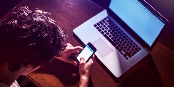 Man sitting in front of a laptop while looking at his mobile phone